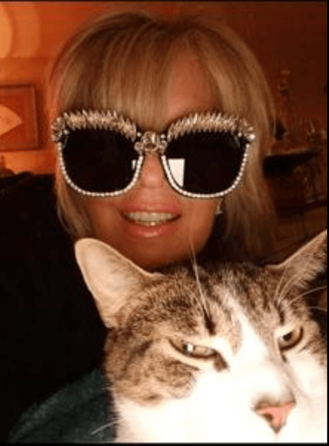 Blonde woman in oversized, jeweled sunglasses smiles next to a grumpy-looking tabby cat.